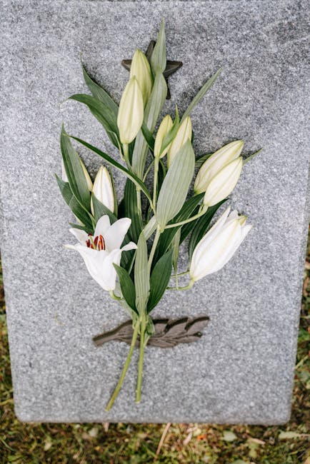 A floral tribute consisting of white lilies and white lilies with green stems and leaves, arranged on a grey stone memorial marker. The lilies are fresh with smooth, slightly curved petals, some in bu