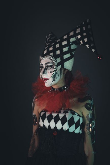 A performer dressed as a clown with elaborate face makeup featuring black and white geometric patterns, exaggerated dark eye makeup, and red lipstick. She is wearing a tall, checkered jester hat in bl