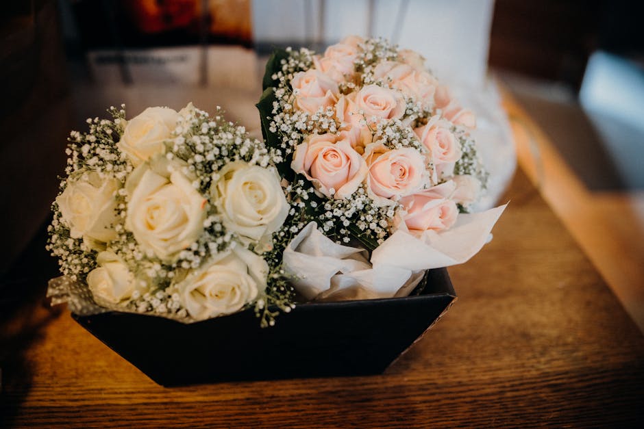 A black rectangular floral arrangement containing two bouquets of fresh roses, one with creamy white roses and the other with soft pink roses, each complemented by delicate sprigs of baby's breath. Th