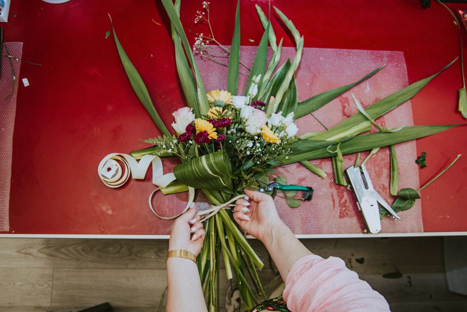 A close-up view of a florist's hands tying a ribbon around a fresh bouquet of flowers on a red work surface. The bouquet features a mix of yellow chrysanthemums, white roses, and small purple flowers,