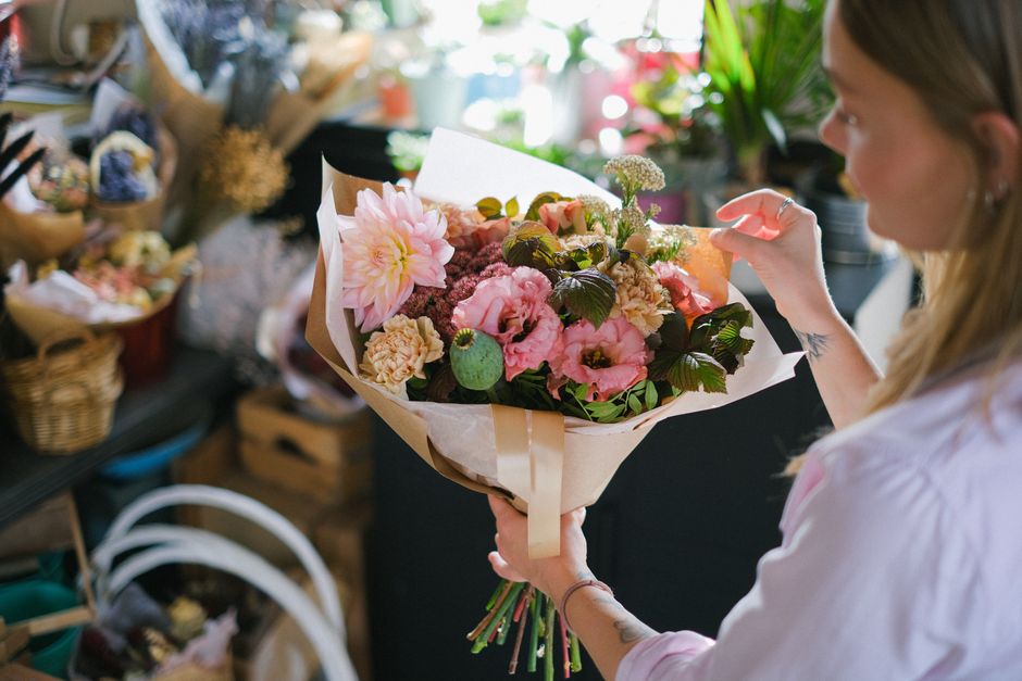 A woman holding a vibrant floral bouquet inside a shop, with the arrangement featuring pink dahlias, light pink lisianthus, dark green leaves, and small white filler flowers, wrapped in beige and whit