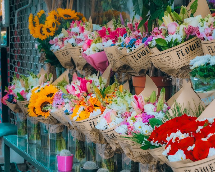 A vibrant display of freshly arranged flower bouquets featuring a variety of blooms including bright yellow sunflowers, pink and white lilies, delicate white and pink roses, and bold red flowers. Each