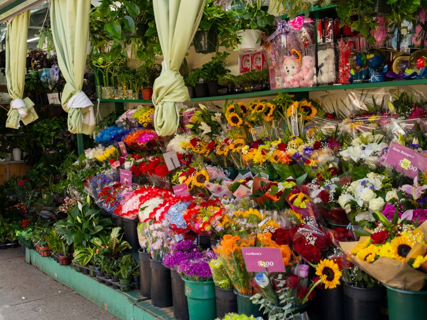 A vibrant outdoor floral display at a florist shop showcasing a variety of fresh flower bouquets arranged on tiered shelves and in buckets. The arrangement includes sunflowers, lilies, carnations, ros
